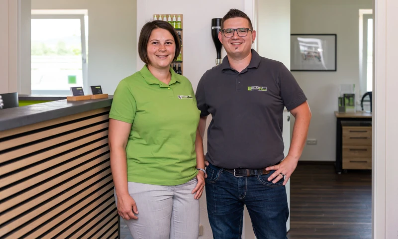 Zwei Mitarbeiter stehen freundlich lächelnd in einem modernen Büro mit Empfangstresen.