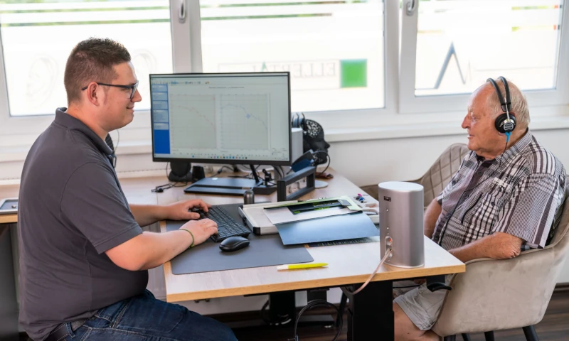 Hörtest beim Akustiker: Älterer Mann mit Kopfhörern, Experte analysiert Audiogramm am PC.